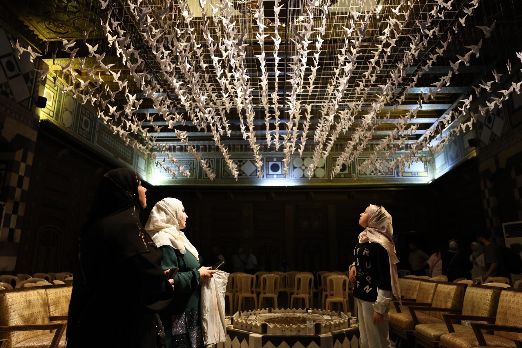 Women look up at hundreds of white paper cranes hanging from the ceiling of an ornate hall, above a circle of empty chairs.