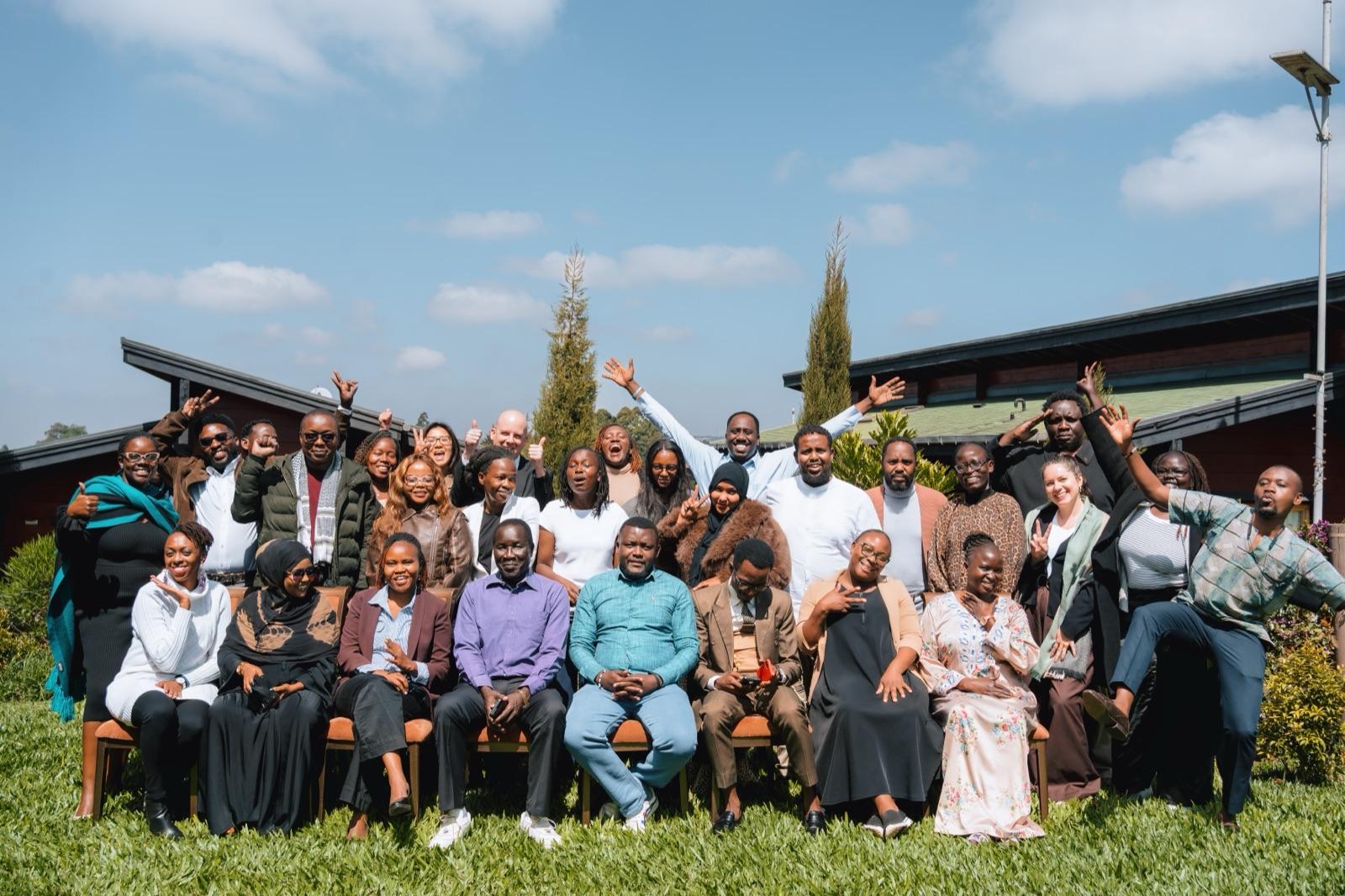 Several dozen refugee leaders and partners pose together on a lawn, many with their arms raised.
