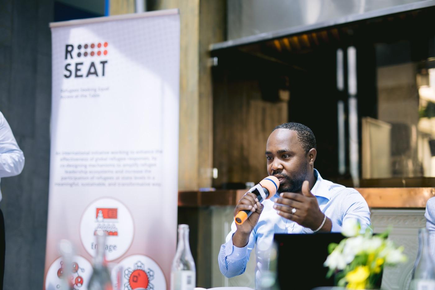 A refugee expert speaks into a microphone at a conference table beside the R-SEAT banner.
