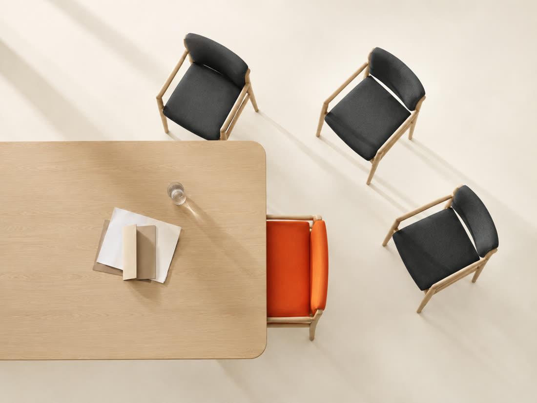 A meeting table seen from above — three chairs pushed back and angled away, one orange chair pulled fully in and squared to the edge
